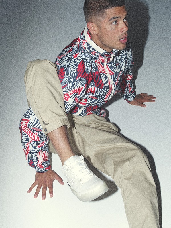 Young man crouching in a red patterned zip jacket, khaki trousers, and white sneakers against a light grey studio background.