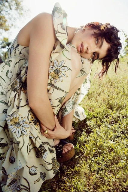 Young woman with dark hair and olive skin poses in a floral print dress in a grassy field, looking at the camera with a neutral expression.