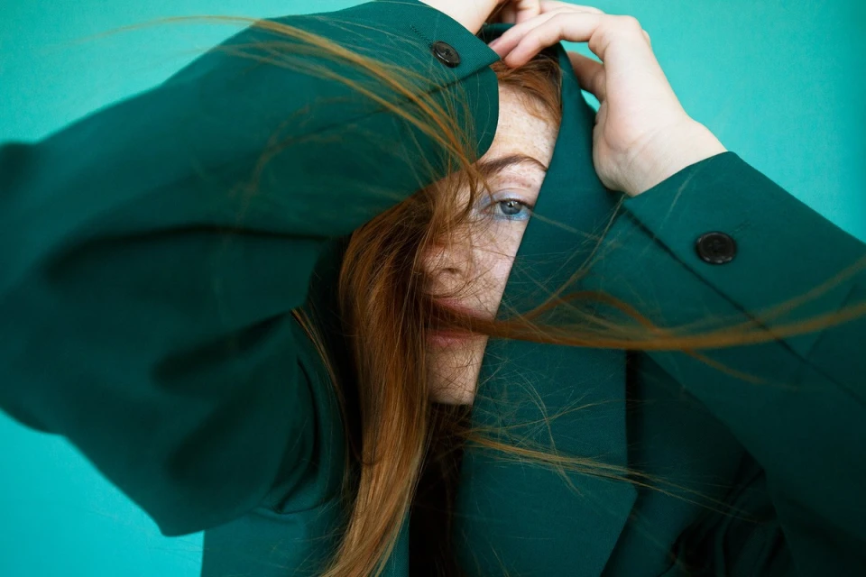 Close-up of a person with red hair wearing a green blazer against a teal background, with the hands holding the jacket around the head.