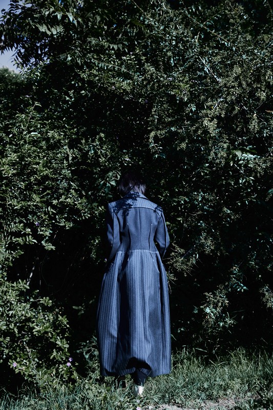 Adult female with dark hair from the back wearing a long blue striped coat in a nature setting with green trees and grass.