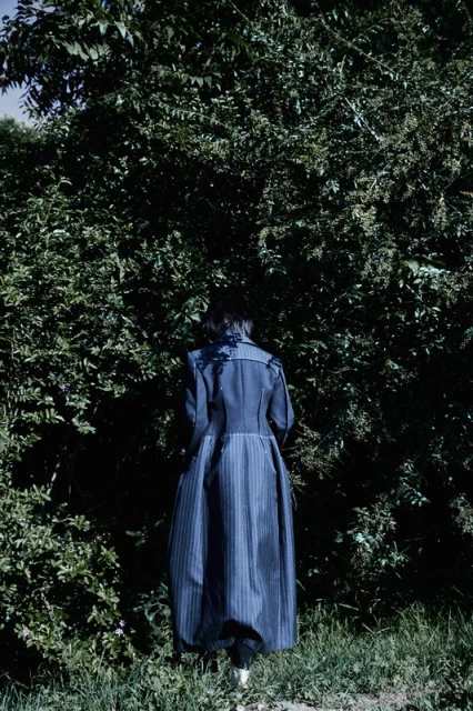 Adult female with dark hair from the back wearing a long blue striped coat in a nature setting with green trees and grass.