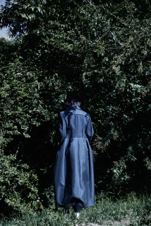 Adult female with dark hair from the back wearing a long blue striped coat in a nature setting with green trees and grass.