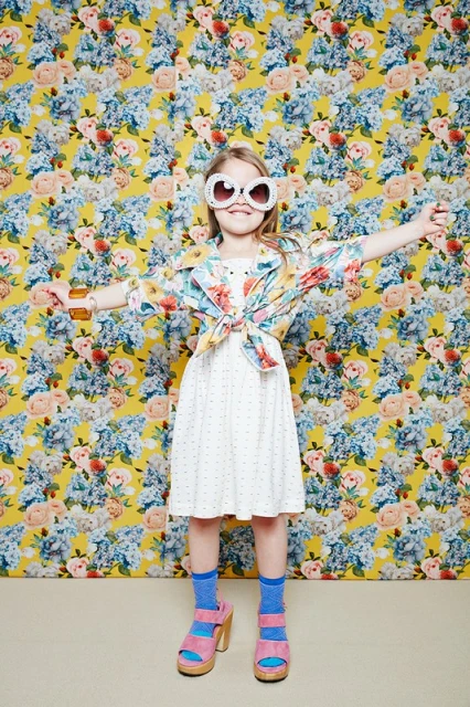 A young girl with outstretched arms poses in front of a floral backdrop, wearing oversized sunglasses, a polka dot dress and pink platform sandals.