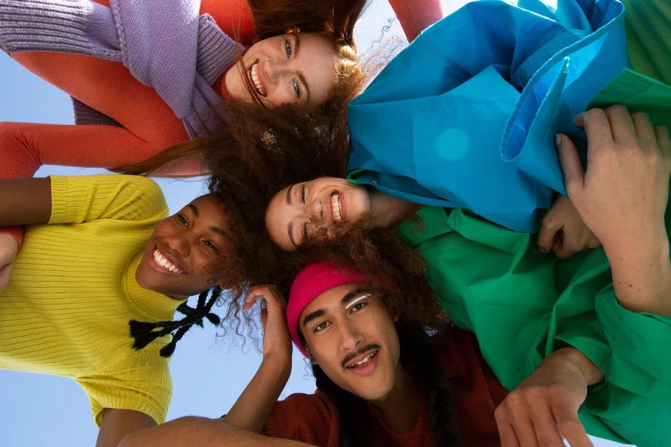 Group of five smiling young adult diverse people in colorful clothing looking down at camera with a bright blue sky in the background.