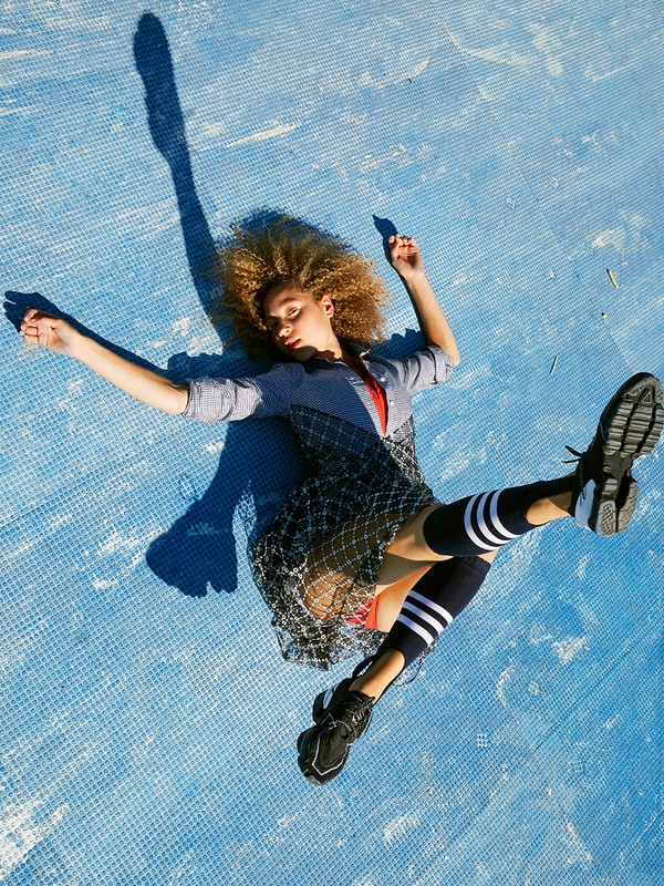 A young adult woman with curly blonde hair lies on a light blue tile-patterned surface, wearing a gray top, black dress, black socks and shoes.
