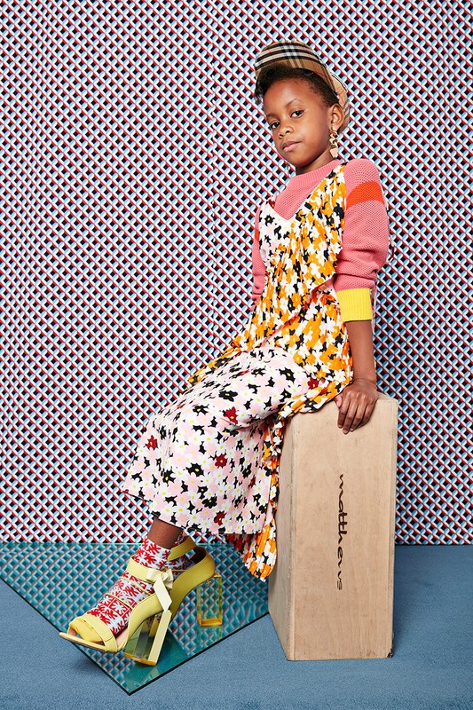 A young girl with a cap sits on a wooden box in a studio with a patterned background, wearing a floral dress, patterned socks, and yellow high heels.