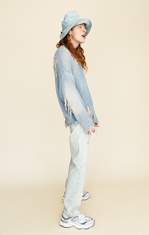 Full-length studio shot of a person wearing a blue bucket hat, ripped sweater, and jeans, posing against a neutral background.