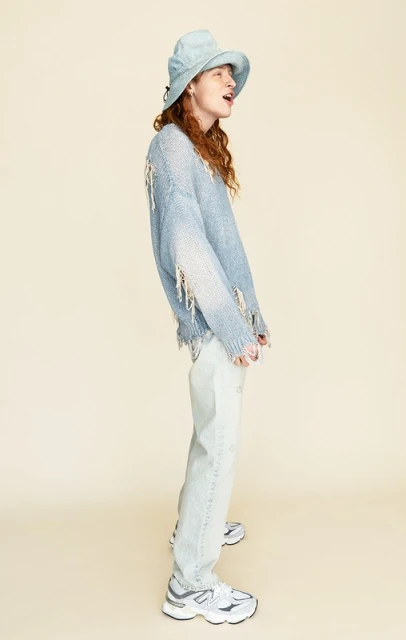 Full-length studio shot of a person wearing a blue bucket hat, ripped sweater, and jeans, posing against a neutral background.