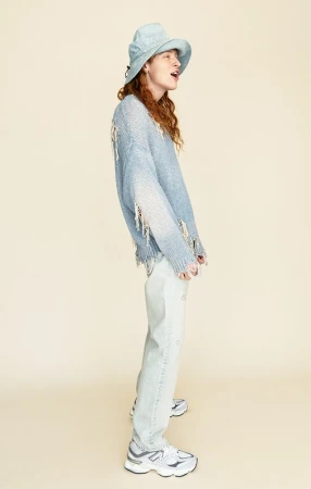 Full-length studio shot of a person wearing a blue bucket hat, ripped sweater, and jeans, posing against a neutral background.