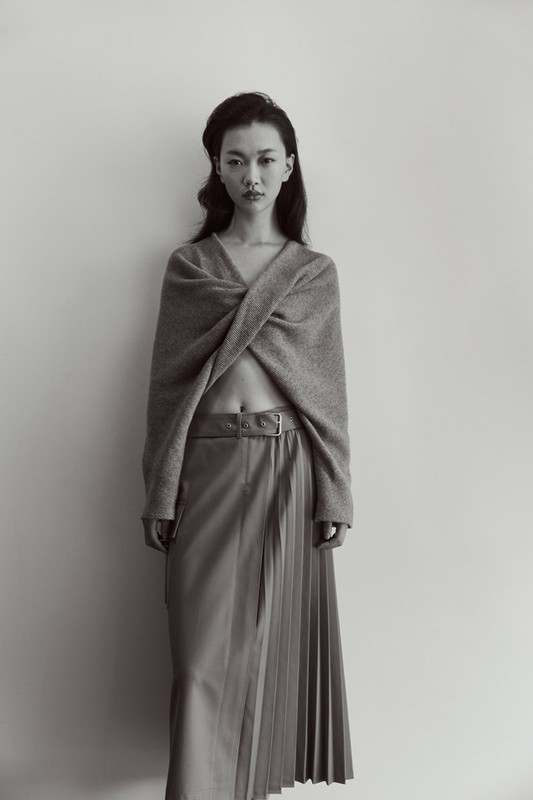 Portrait of a young woman in grey knitwear and a pleated skirt against a white background. Fashion shot, studio lighting.