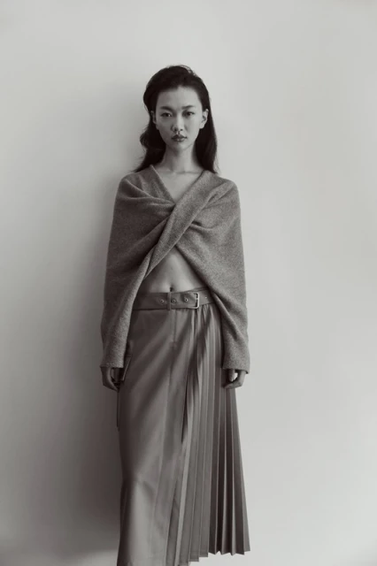 Portrait of a young woman in grey knitwear and a pleated skirt against a white background. Fashion shot, studio lighting.