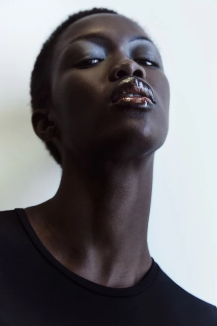 Close-up portrait of a dark-skinned woman with glossy lips wearing a black top, set against a clean white background.