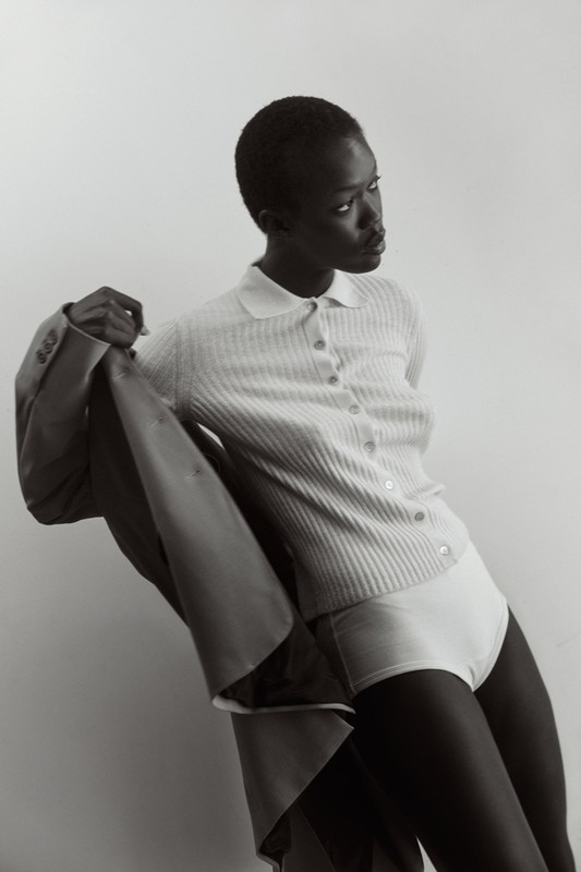 Elegant portrait of a slender dark-skinned woman in a ribbed shirt and briefs, holding a suit jacket in a neutral studio setting.