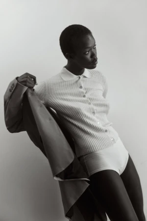 Elegant portrait of a slender dark-skinned woman in a ribbed shirt and briefs, holding a suit jacket in a neutral studio setting.