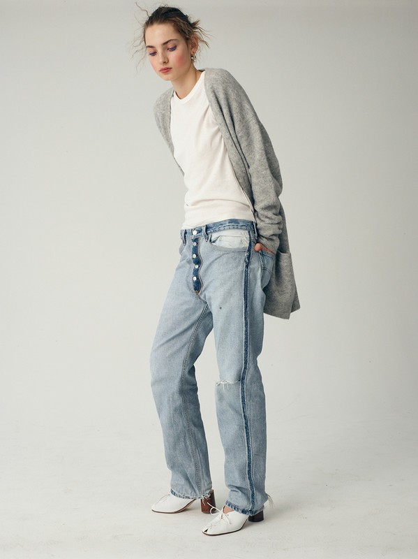 Young woman in casual fashion stands in a studio: white t-shirt, grey cardigan, deconstructed jeans, white heels against a neutral backdrop.