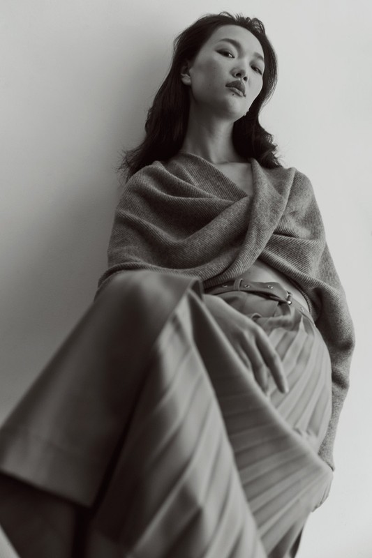 Monochrome portrait of an Asian woman in a shawl and pleated skirt against a neutral wall, capturing understated elegance and style.