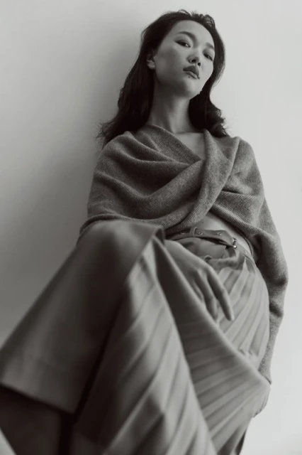 Monochrome portrait of an Asian woman in a shawl and pleated skirt against a neutral wall, capturing understated elegance and style.