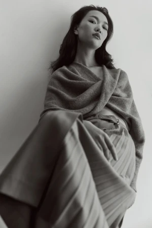 Monochrome portrait of an Asian woman in a shawl and pleated skirt against a neutral wall, capturing understated elegance and style.