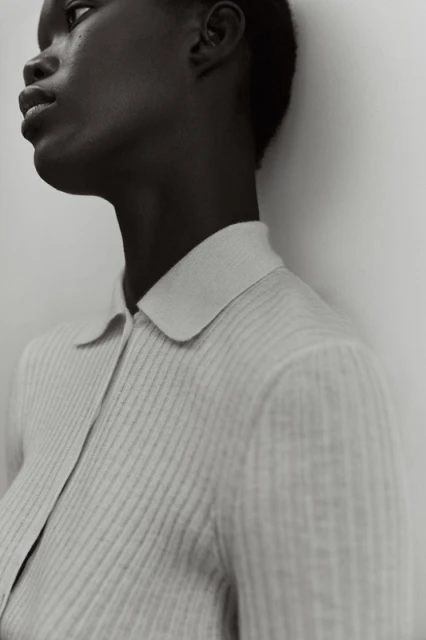 Close-up of a Black female model in a light gray ribbed collared shirt against a light gray background, highlighting the shirt's texture.