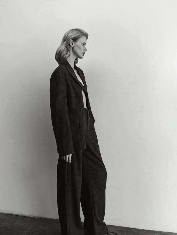 A woman in a black blazer and wide-leg pants poses in profile against a white wall, showcasing fashion-forward editorial style.