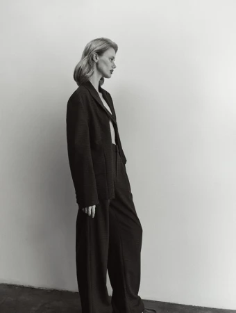 A woman in a black blazer and wide-leg pants poses in profile against a white wall, showcasing fashion-forward editorial style.