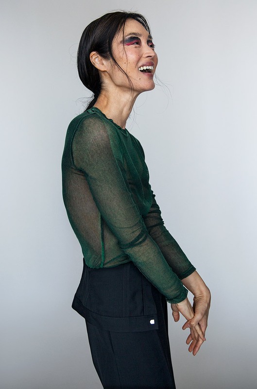 Profile view of smiling woman wearing a sheer dark green long-sleeve top and black trousers against a neutral background.