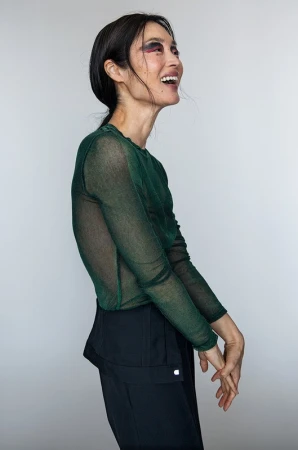 Profile view of smiling woman wearing a sheer dark green long-sleeve top and black trousers against a neutral background.