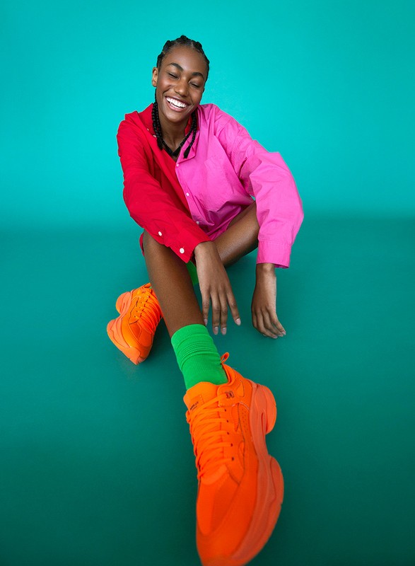 Smiling young African-descent woman in a pink and red shirt, green socks, and orange sneakers against a teal backdrop.