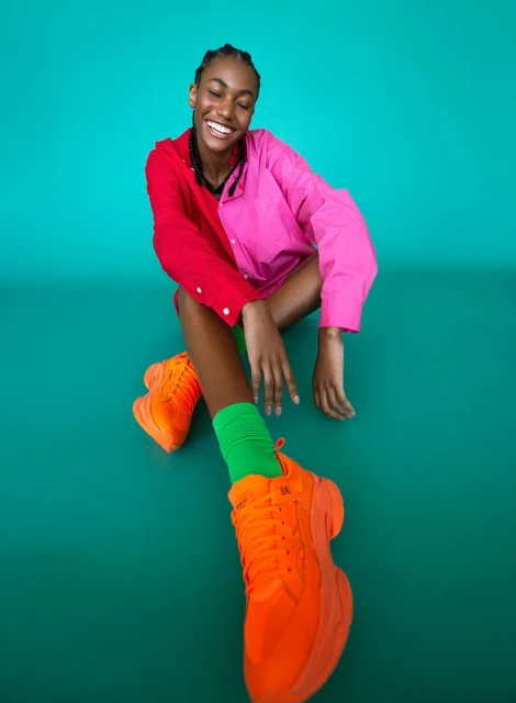 Smiling young African-descent woman in a pink and red shirt, green socks, and orange sneakers against a teal backdrop.