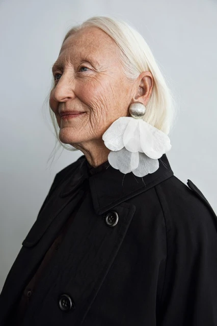 Elegant older woman in a black trench coat with large earrings in a side-profile portrait against a neutral backdrop.