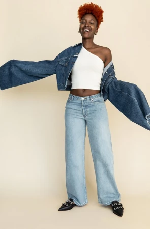 Black woman with curly orange hair wearing a white one-shoulder top, denim jacket, and light-wash jeans against a seamless backdrop