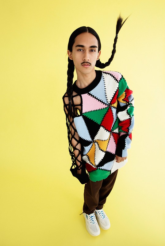 Full body portrait of a young adult male wearing a patterned sweater, sweatpants, and sneakers against a solid yellow background.