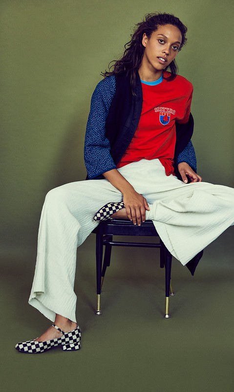 Woman with long dark hair seated on a dark chair wearing a red t-shirt, patterned blue sleeves, and wide white trousers against an olive green background.
