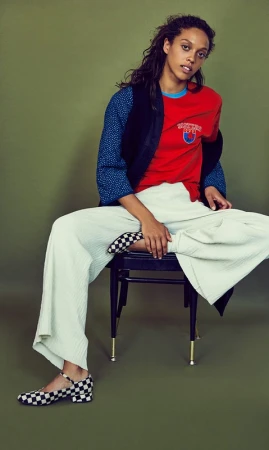 Woman with long dark hair seated on a dark chair wearing a red t-shirt, patterned blue sleeves, and wide white trousers against an olive green background.