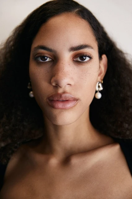 Close-up portrait of an adult woman with dark curly hair and natural makeup wearing drop earrings against a neutral background.