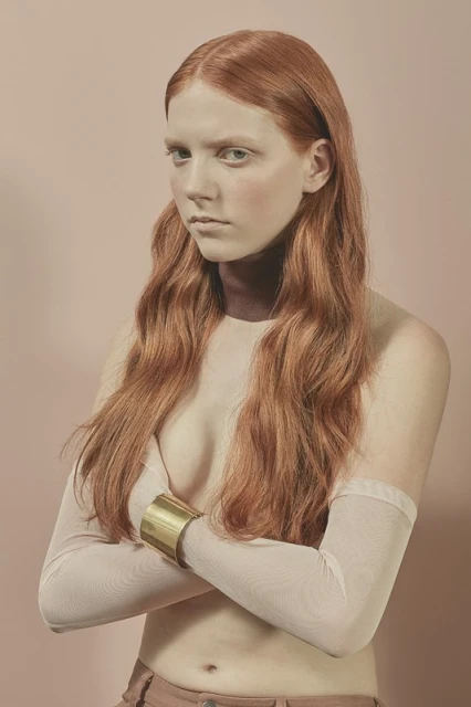 Portrait of a red-haired, pale-skinned woman with arms crossed, wearing a gold cuff bracelet and sheer sleeves against a pink backdrop.