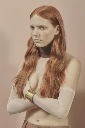 Portrait of a red-haired, pale-skinned woman with arms crossed, wearing a gold cuff bracelet and sheer sleeves against a pink backdrop.