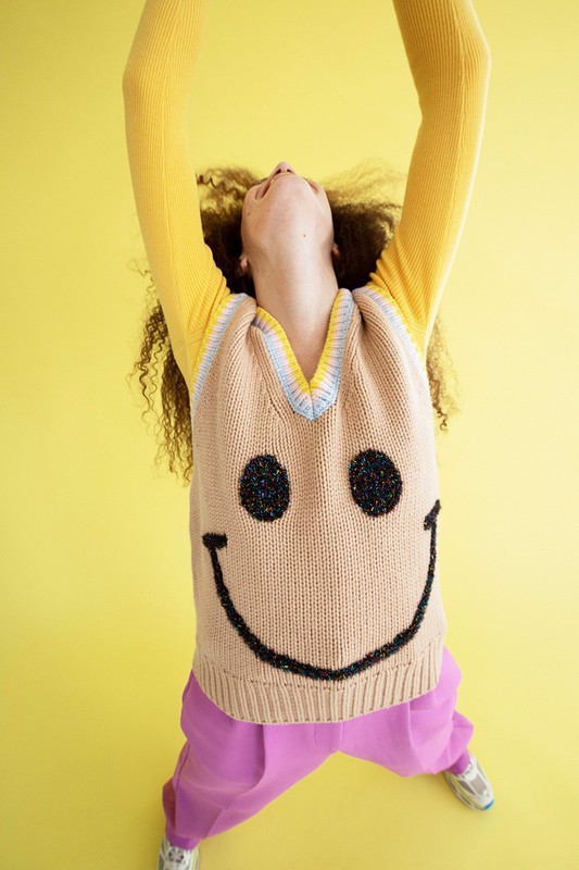 Woman wearing a smile face vest in a high angle shot on a yellow backdrop with raised arms, showcasing fashion and positive style.