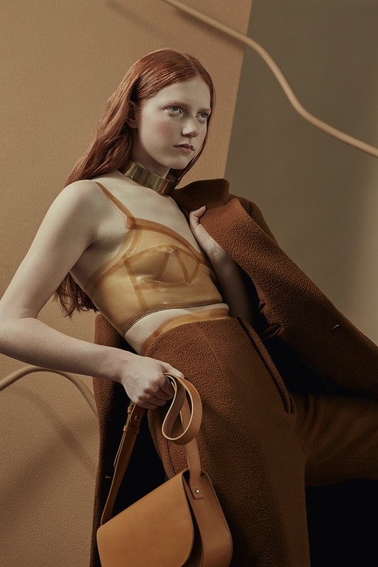 A woman with red hair poses indoors wearing a brown outfit, gold collar, and clear top. She holds a brown handbag with a tan background.