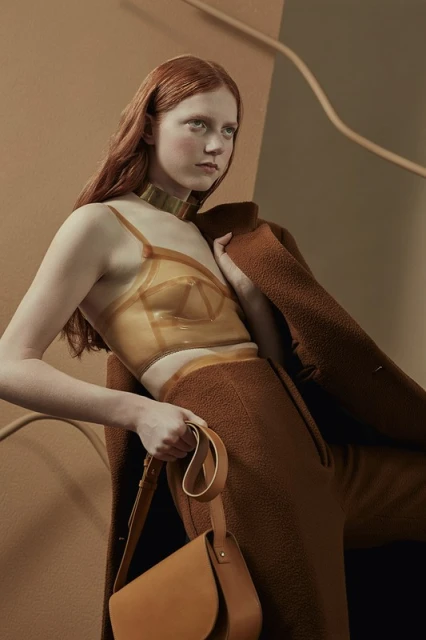 A woman with red hair poses indoors wearing a brown outfit, gold collar, and clear top. She holds a brown handbag with a tan background.