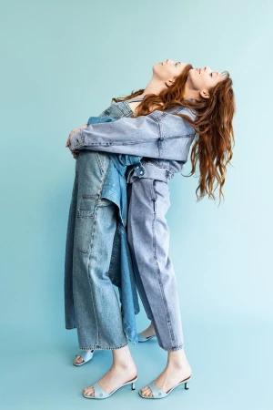 Two red-haired women in matching denim outfits embrace in front of a light blue backdrop. One wears overalls, the other a jacket and pants.