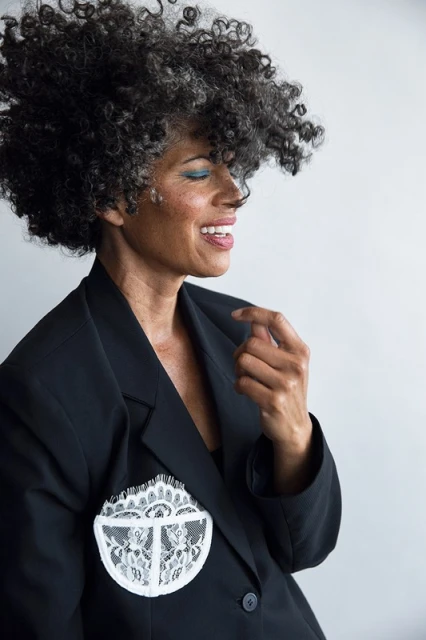 Smiling woman in a black blazer with white lace detail, side profile on a light grey background, with a full head of curly grey hair.
