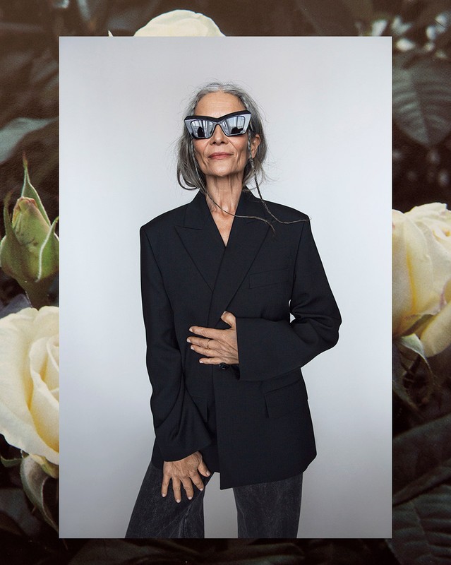 Mature woman in sunglasses and an oversized blazer poses in front of roses, exuding a confident and stylish aesthetic, fashion forward.