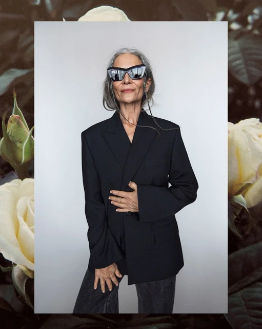 Mature woman in sunglasses and an oversized blazer poses in front of roses, exuding a confident and stylish aesthetic, fashion forward.