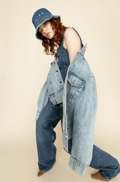 A young woman poses in a denim outfit and bucket hat, complemented by a light blue denim jacket, set against a cream-colored backdrop.