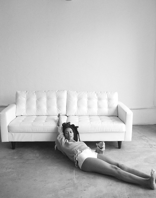 Black and white portrait of a woman with locs lying on a concrete floor in front of a white couch and white wall in an indoor setting.