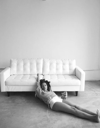 Black and white portrait of a woman with locs lying on a concrete floor in front of a white couch and white wall in an indoor setting.