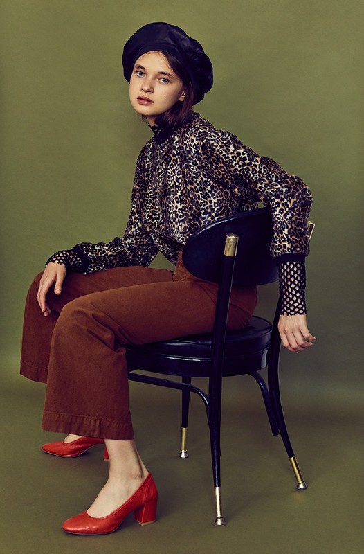 Portrait of a young woman in a black beret and animal-print blouse sitting on a chair in a studio against a green background.