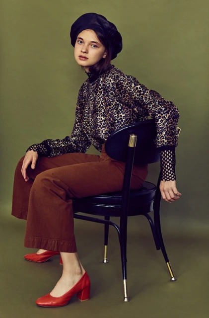 Portrait of a young woman in a black beret and animal-print blouse sitting on a chair in a studio against a green background.