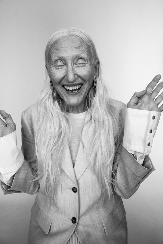 Black and white portrait of an elderly woman with long white hair, closed eyes, and a big smile, wearing a blazer in a bright studio.